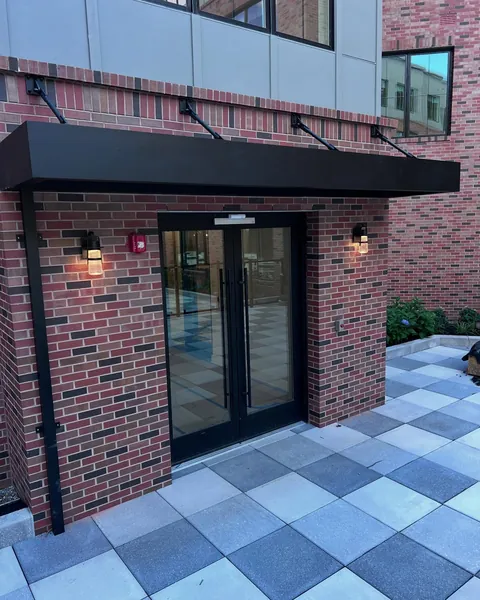 Black aluminum canopy with tie-rod supports above glass double doors on a brick building.