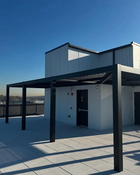 Black louvered pergola on a rooftop deck beside a white building.