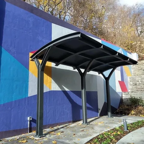 Curved dark shade structure with three support posts in front of a colorful wall mural.